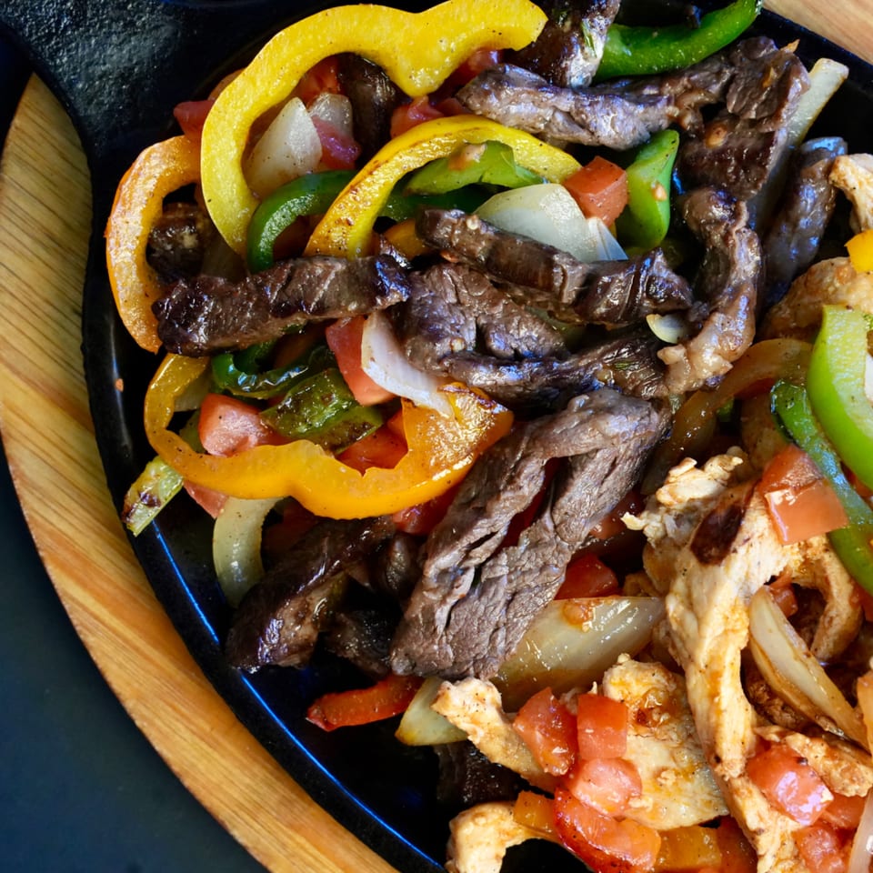 Sizzling fajitas with steak, chicken, colorful bell peppers, and onions on a black skillet.